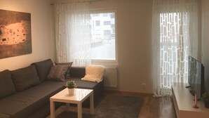 Una sala de estar con un sofá, una mesa de centro con una planta, un cuadro en la pared y una ventana con persianas.
