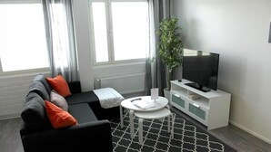 A modern living room with a black sofa, orange cushions, a white coffee table, a black and white patterned rug, a white TV stand, and a potted plant. - Kotimaailma Apts Joensuu Koskikatu 11 (Joensuu)