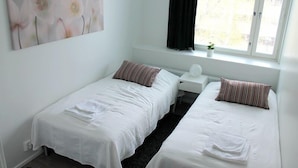 A room with two single beds, a small white nightstand, a window with black curtains, and a wall-mounted artwork of flowers. - Kotimaailma Apts Joensuu Koskikatu 11 (Joensuu)