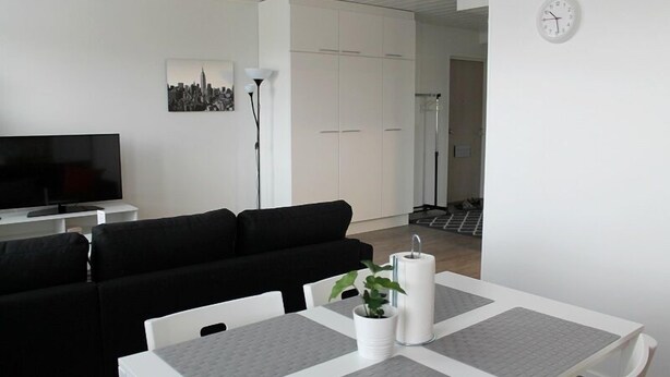 A modern living room with a black sofa, a white dining table with gray placemats, a TV, and a cityscape painting on the wall. - Kotimaailma Apts Joensuu Koskikatu 11 (Joensuu)