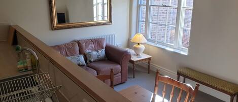 Una sala de estar con un sofá, una mesa con una lámpara, una silla de madera, un espejo grande y una ventana con vista a un edificio de ladrillo.