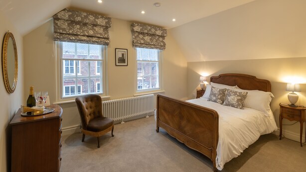 A bedroom with a wooden bed, two windows with floral valances, a radiator, a wooden dresser, a chair, and a bedside table with a lamp. - 71 Micklegate - Apartment 2 (York)