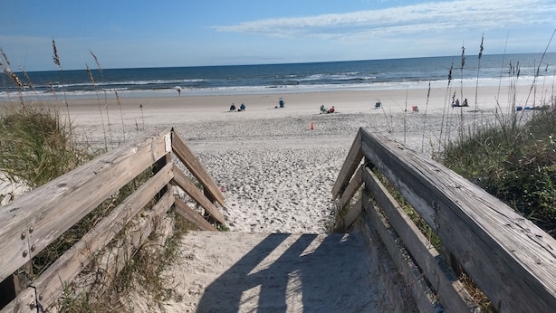 Une plage de sable avec des passerelles en bois menant à l’eau, où l’on peut voir des gens assis et se promenant le long du rivage.