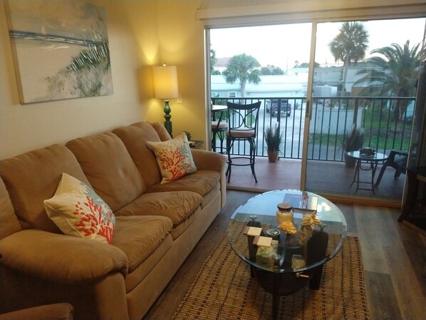 A living room with a brown sofa, two decorative pillows, a glass coffee table with various items, a lamp, and a view of a balcony with chairs and a table.