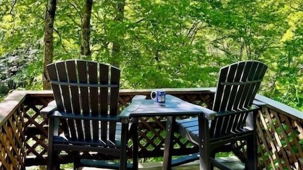 Eine Holzterrasse mit zwei Adirondack-Stühlen und einem Tisch, umgeben von üppigem Grün.