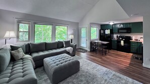 Ein modernes Wohnzimmer mit einem grauen Ecksofa, einem passenden Hocker und einem Essbereich mit kleinem Tisch und Stühlen. Durch eine offene Tür ist eine Küche mit grünen Schränken und einem schwarzen Kühlschrank zu sehen.
