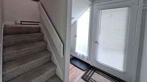 Eine Treppe mit Teppichstufen führt zu einer Zwischenebene, eine Tür mit Jalousien und eine Wandleuchte.
