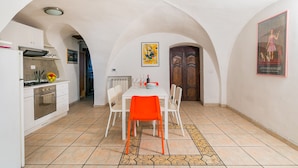 Un coin repas avec une table blanche, des chaises orange et un sol carrelé. On y trouve une cuisine avec des placards blancs et un fourneau. Une porte en bois et un tableau encadré sont visibles sur les murs.