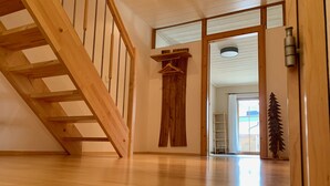 Un escalier en bois menant à un deuxième étage, un porte-manteau en bois, un petit sapin de Noël en bois et une vue sur un balcon avec une balustrade.