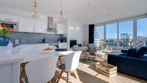 A modern kitchen with white cabinets, a central island, and a dining area with white chairs. There is a blue sofa and a wooden crate in the living area. - Water house "The Nordic House" at Ostseeresort Olpenitz - sun deck & sea view (Olpenitz)
