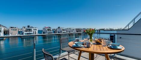 A wooden deck with a round table set for dining, overlooking a marina with boats and buildings in the background.