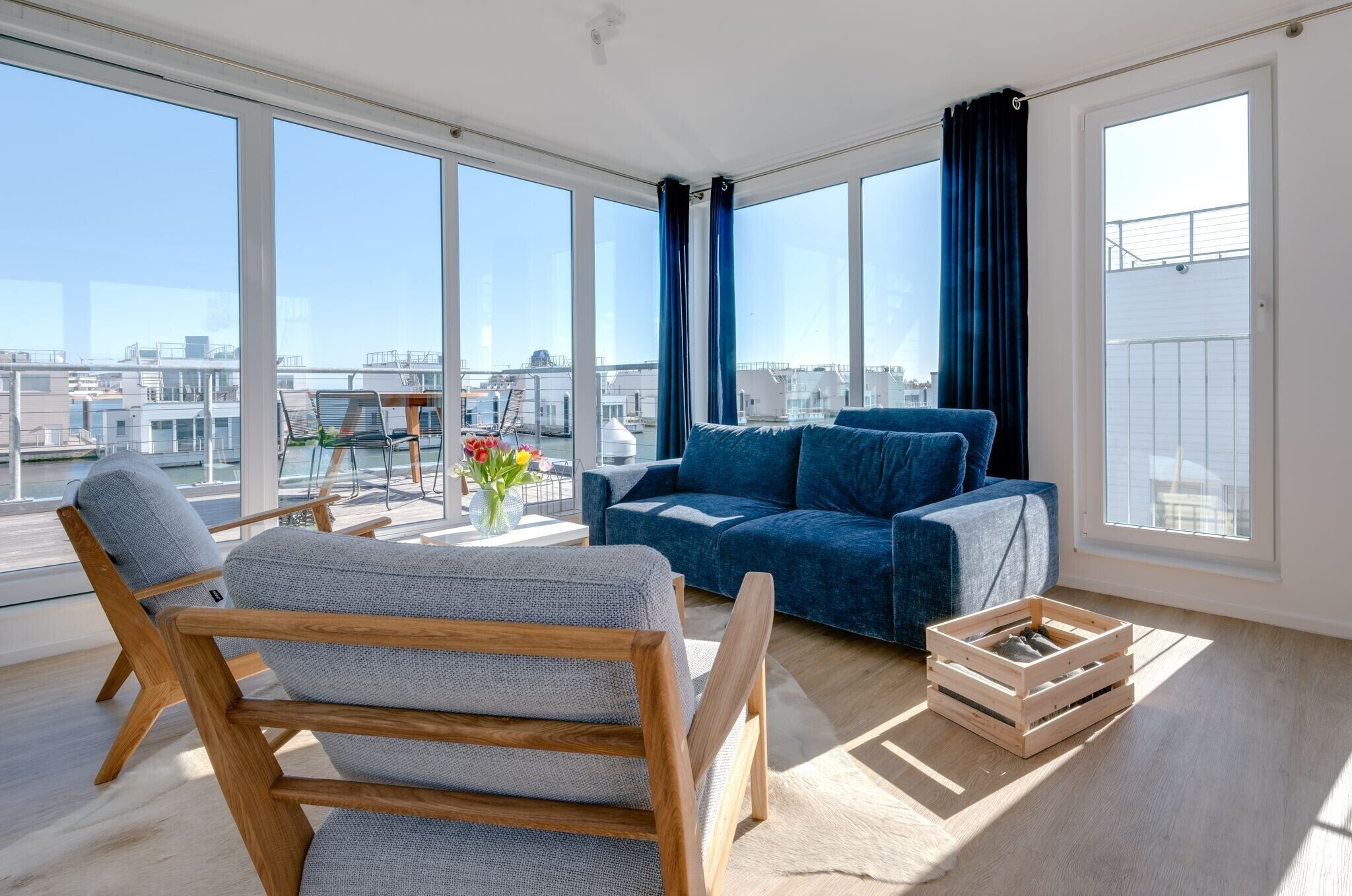 Una sala de estar moderna con ventanas grandes, un sofá azul, una silla de madera y una mesita con flores.