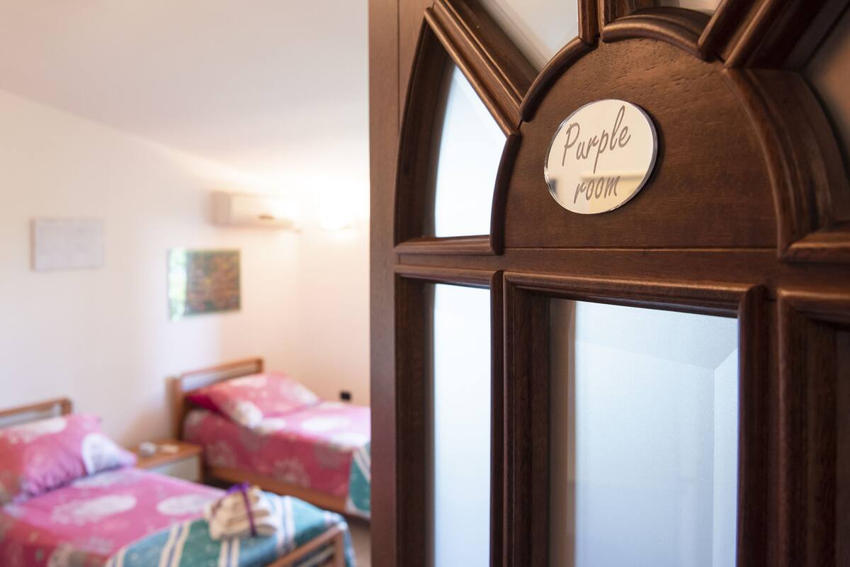 A room with a sign reading "Purple Room" on a wooden door. There are two beds with floral bedding.