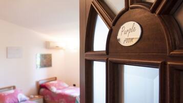 Une pièce dont la porte en bois porte une pancarte indiquant « Chambre violette ». On y trouve deux lits avec une literie à motifs floraux.