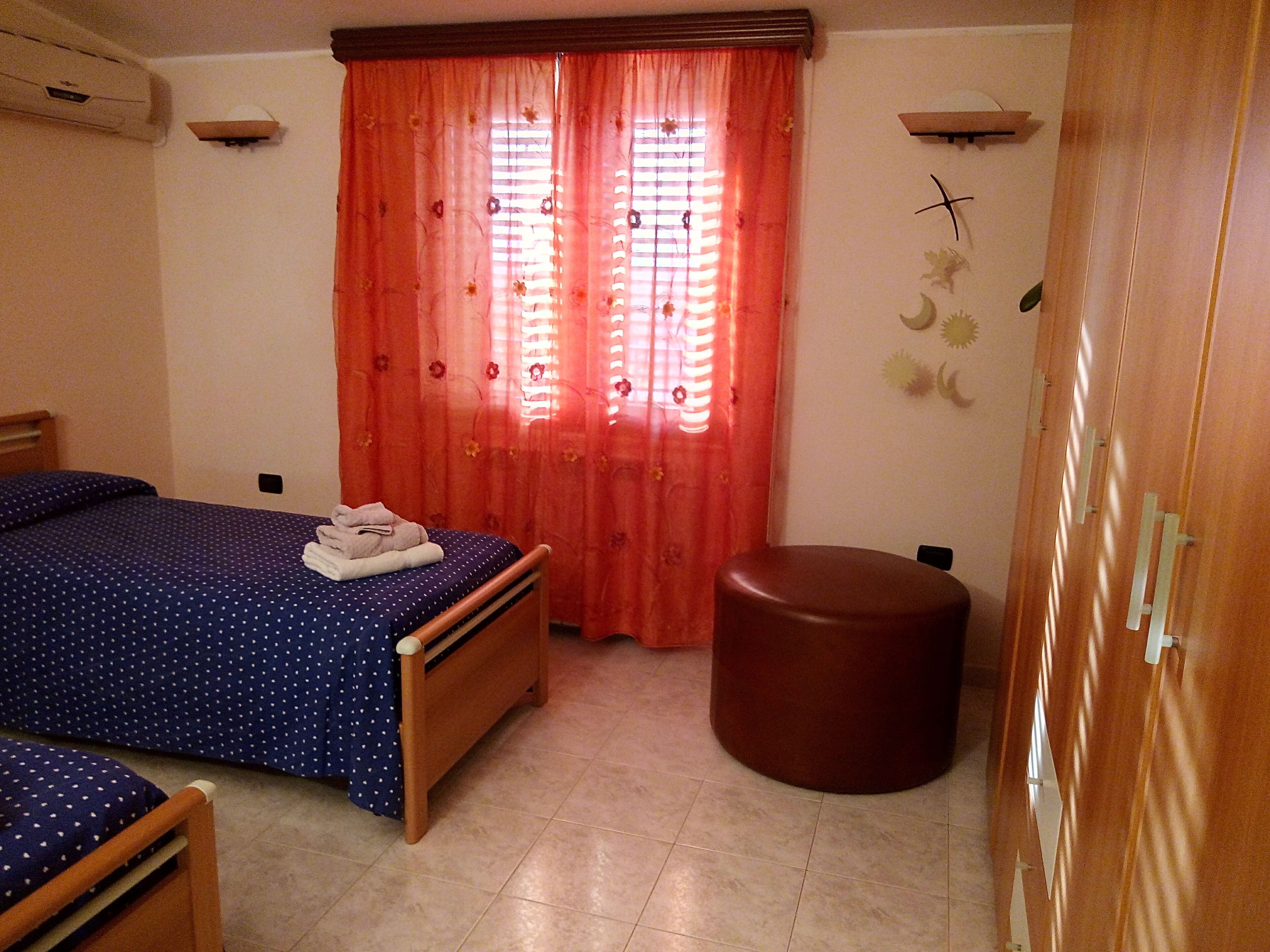 A bedroom with two beds, a wooden wardrobe, a round ottoman, and a window with red curtains.