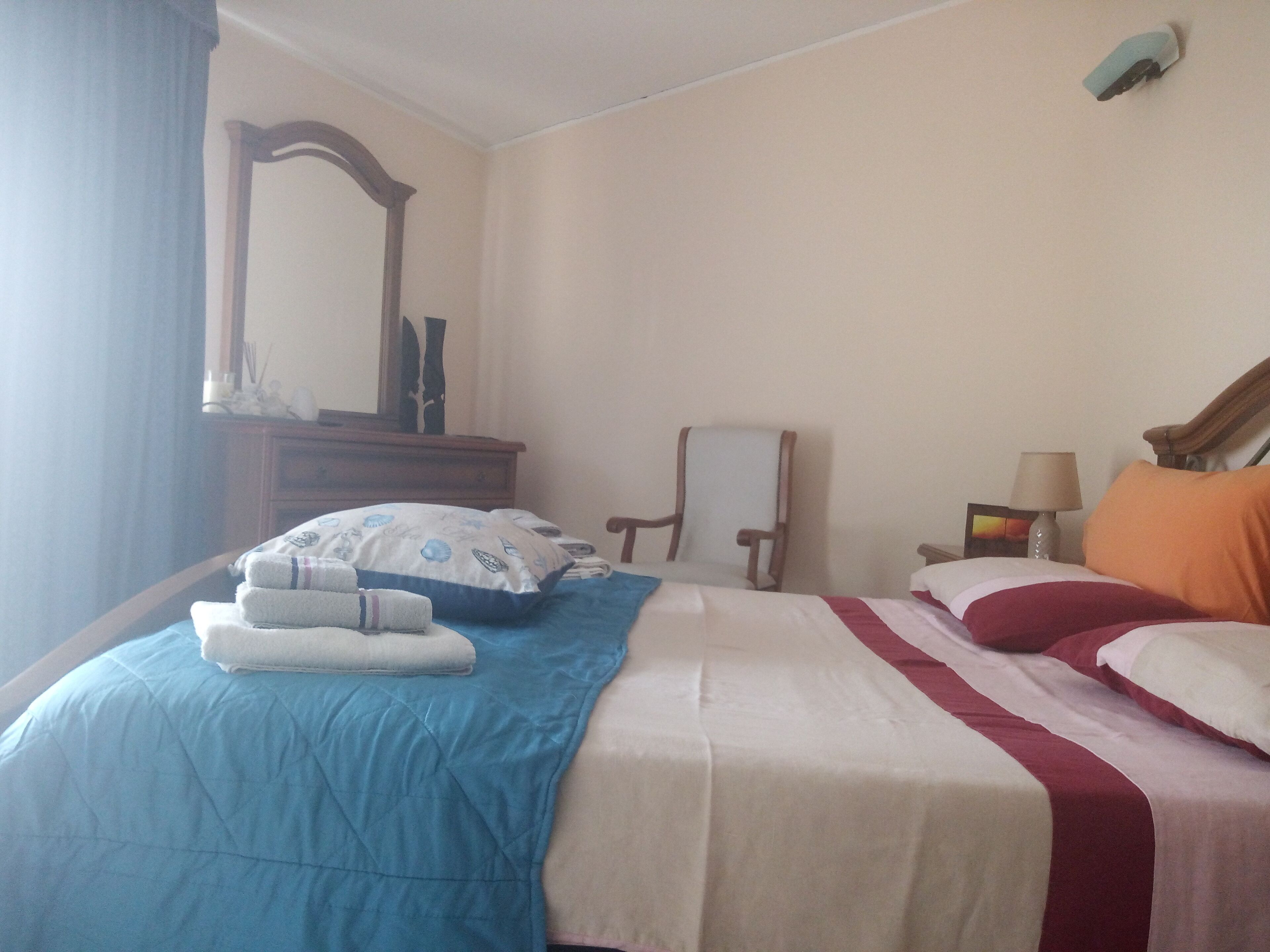 Ein ordentlich gemachtes Bett mit einer blau-weiß gestreiften Tagesdecke, ein Nachttisch mit einer Lampe und ein Stuhl an der Wand.