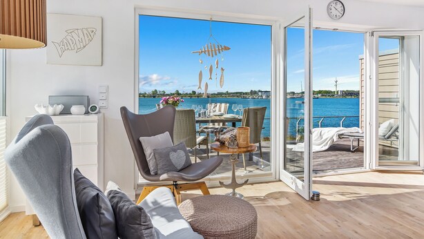 A modern living room with a view of the sea, a dining area with outdoor seating, and a large window with a clock.