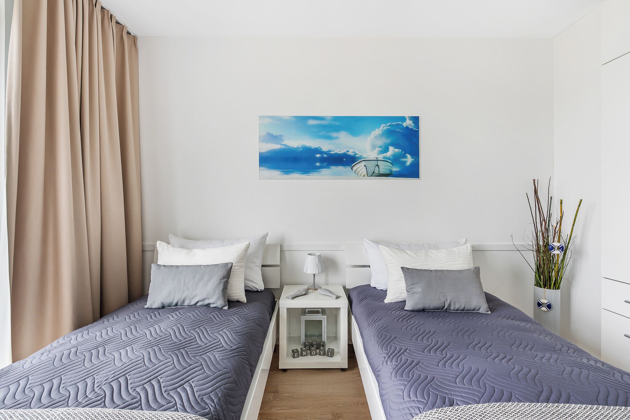 A hotel room with two beds, a nightstand with a lamp, a painting of a boat on water, and a vase with plants.