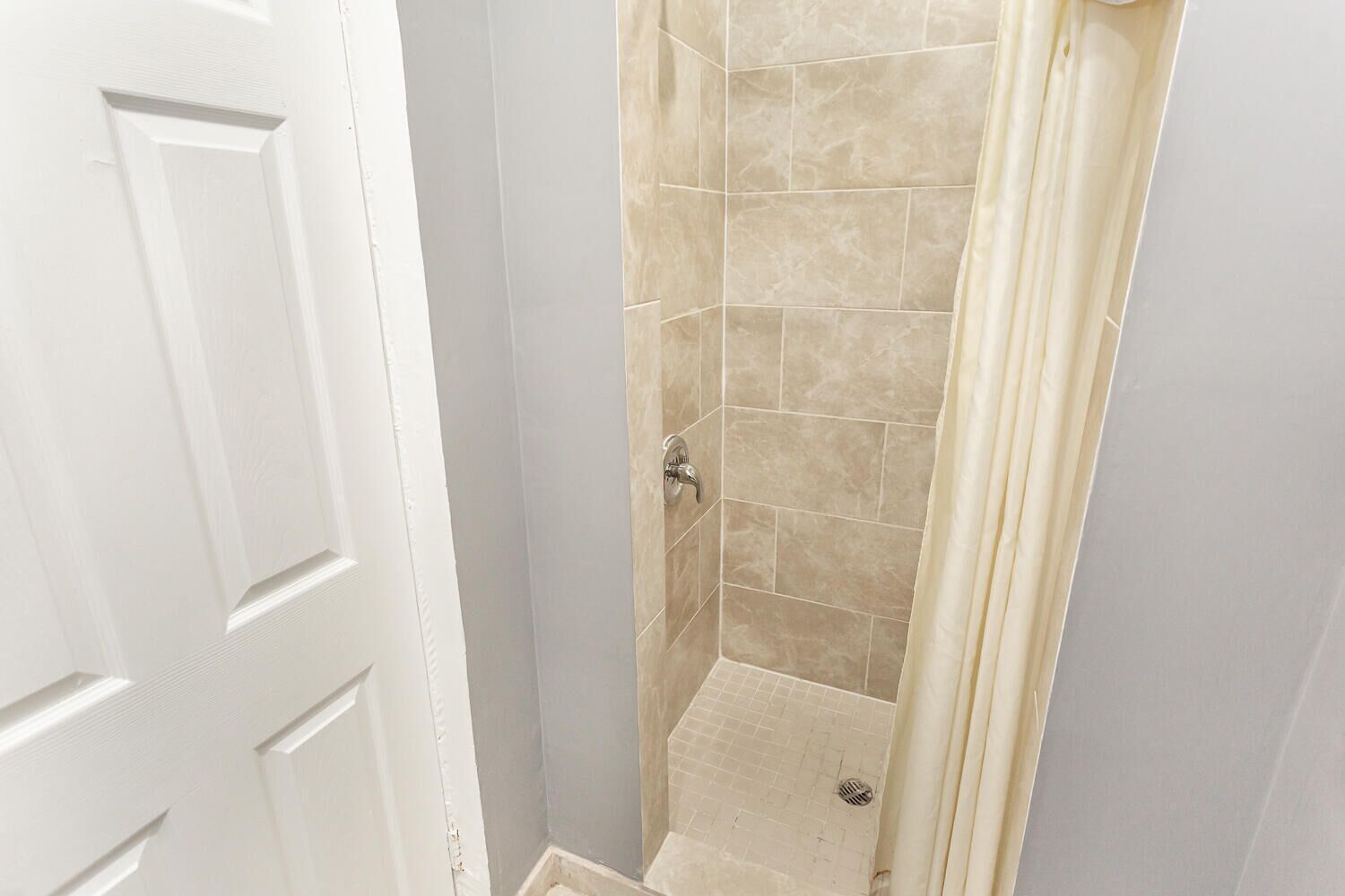 Un baño con azulejos color beige, una cortina de ducha y una puerta blanca.