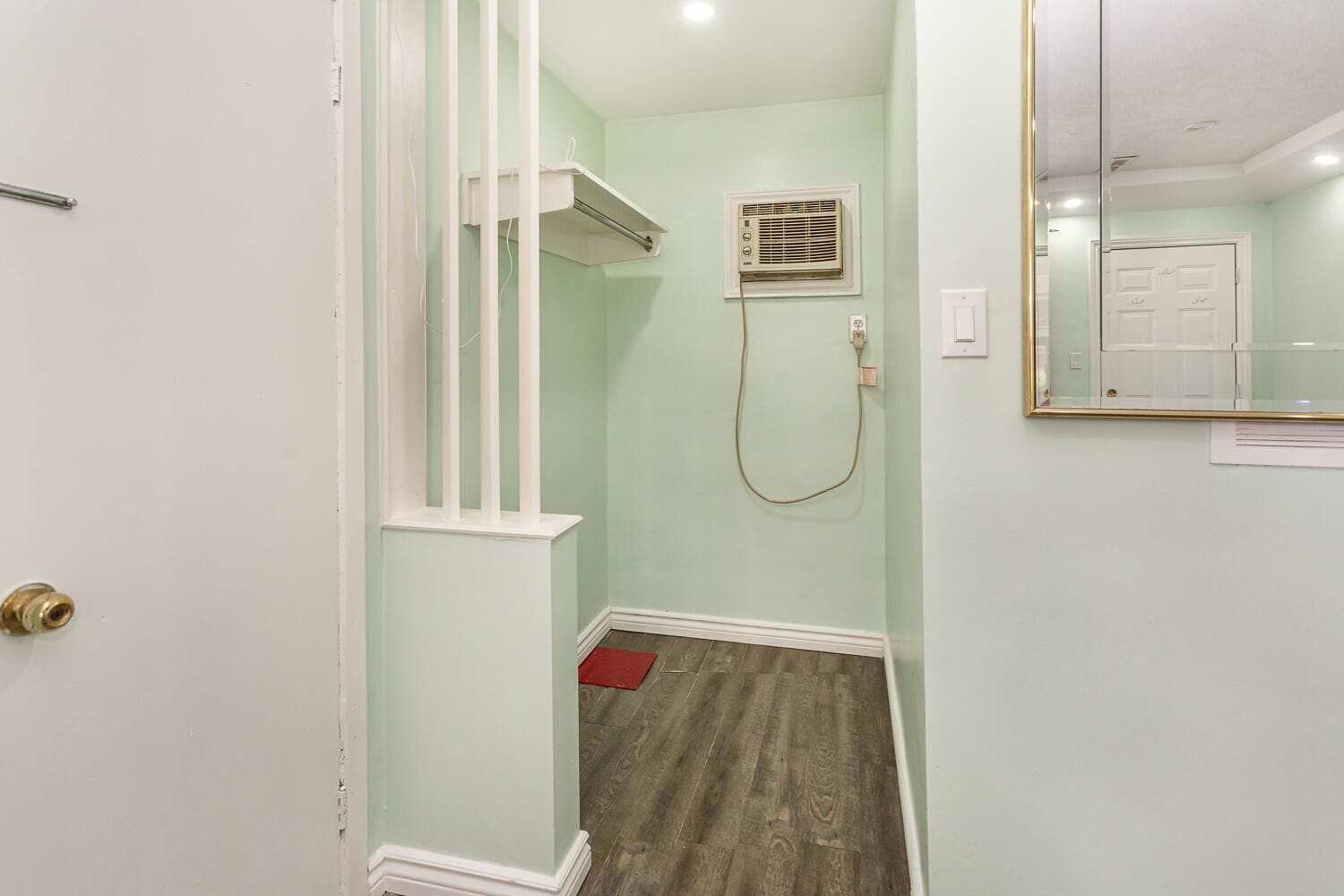 A narrow hallway with a wooden floor, a white door, a small red mat, and a white-framed mirror on the wall.