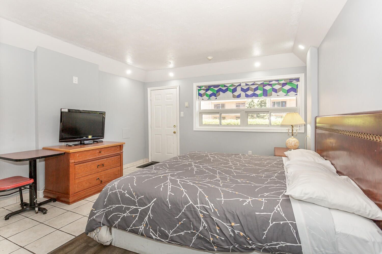 Un dormitorio con cama, escritorio con ordenador, televisor y ventana con vista a edificios.