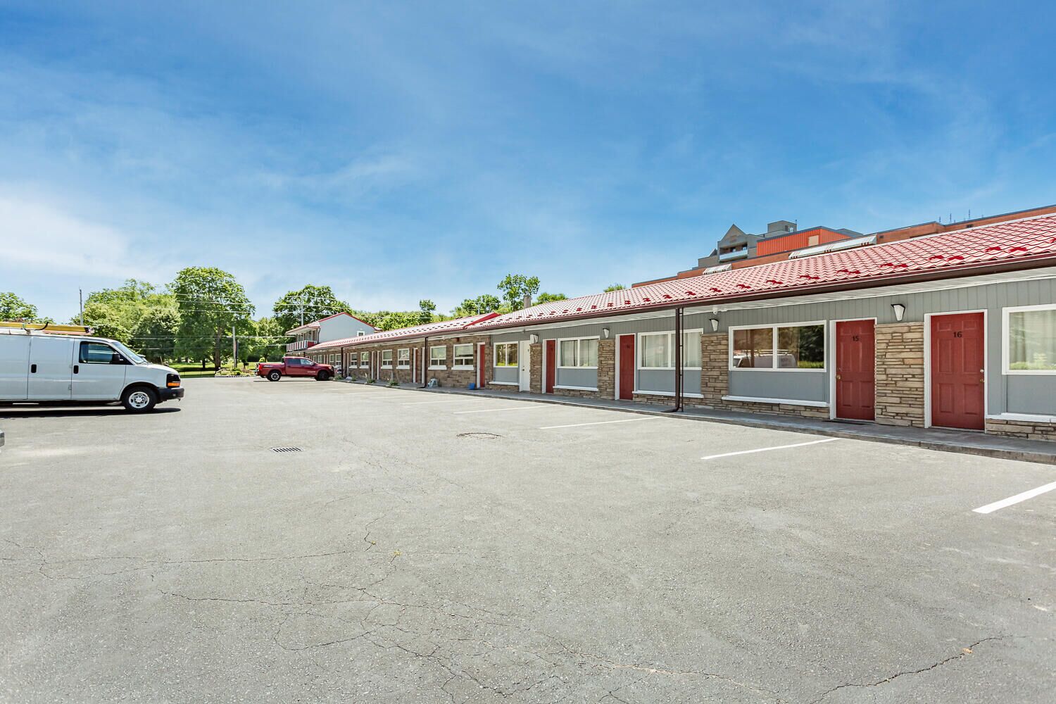 A motel with a row of rooms, each with a red door and a window. There is a white van and a red truck parked in the lot.