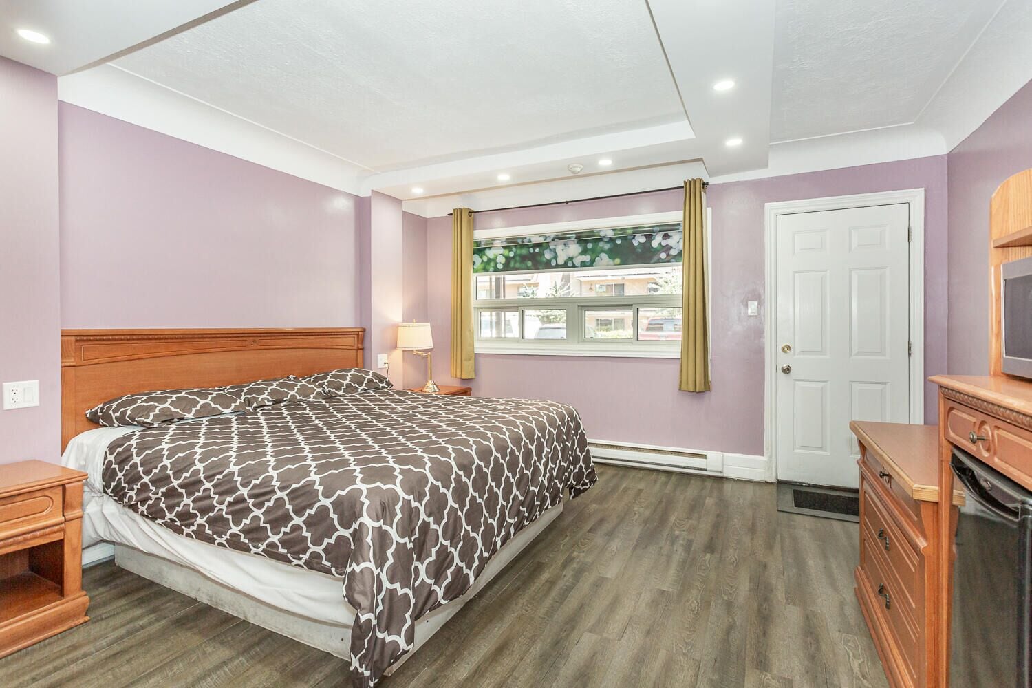 A bedroom with a bed, a nightstand, a dresser, a TV, and a window with a view of buildings.