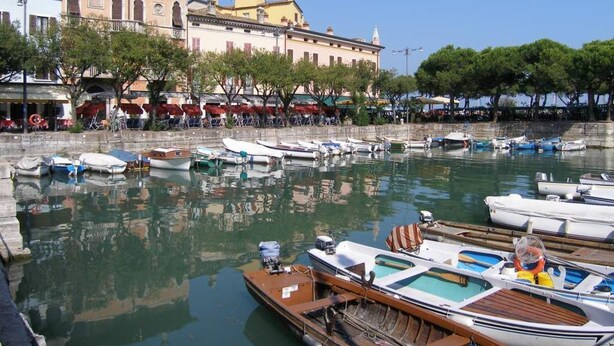 Un puerto deportivo con barcos amarrados, un restaurante frente al agua con terraza, y edificios históricos al fondo.