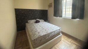 Een kleine, netjes ingerichte slaapkamer met een enkel bed, een hoofdbord met een geometrisch patroon, een raam met gordijnen en een radiator.