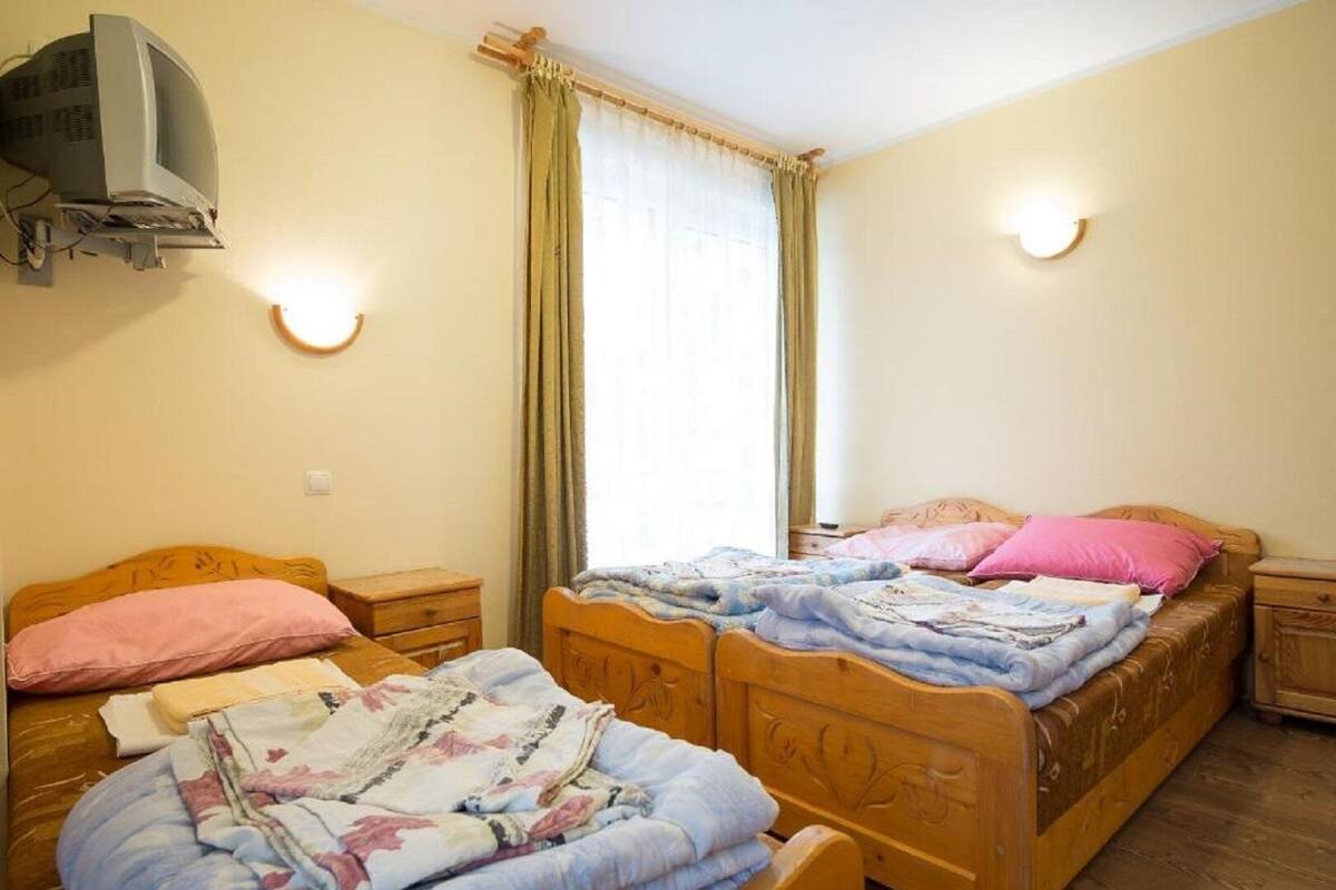 A room with two wooden beds, each with a headboard, a bedside table, and a lamp. There is a window with curtains, a wall-mounted TV, and a wall sconce.