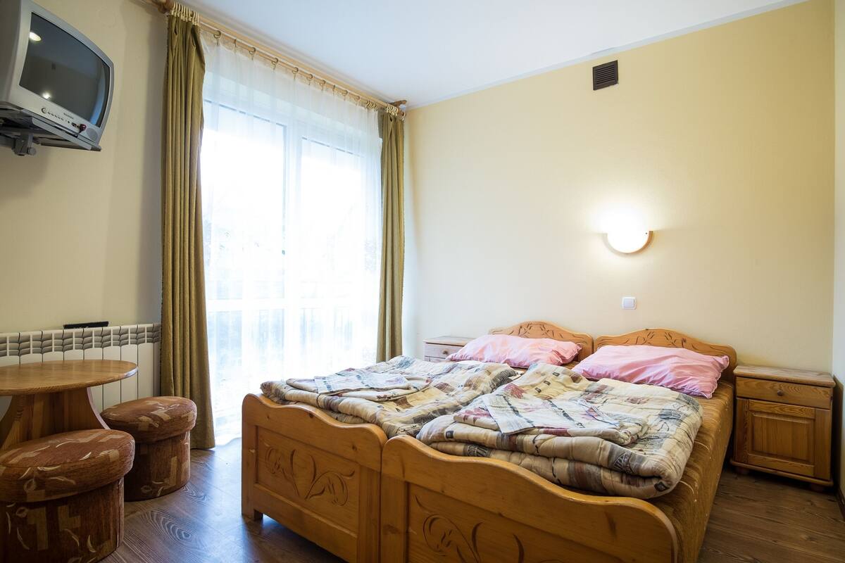 A bedroom with a wooden bed, a bedside table, a small wooden nightstand, a round wooden table, and a television mounted on the wall.