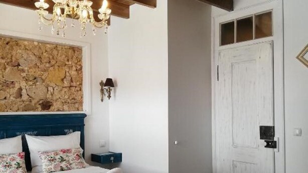 Un dormitorio con techo de madera, una araña, una pared de piedra, una cama con almohadillas florales y una puerta blanca.