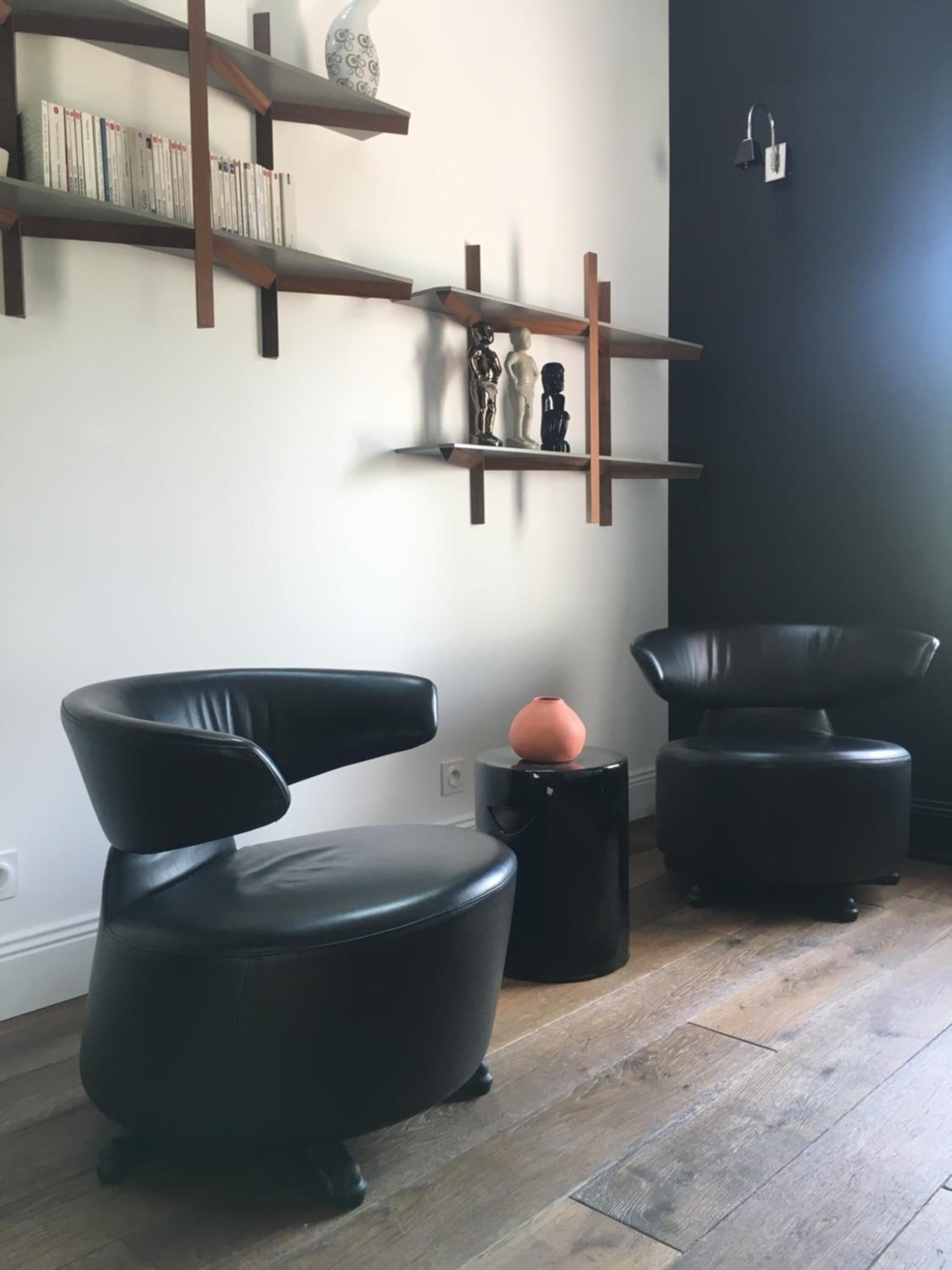 Een moderne woonkamer met een zwarte lederen fauteuil, een rond pouf en een zwarte salontafel. Er staan houten planken met boeken en decoratieve objecten.