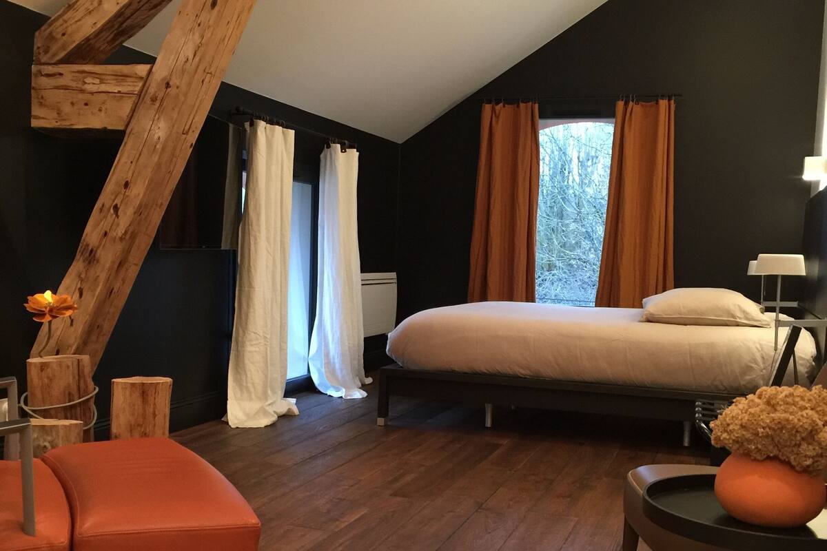 Quarto com teto inclinado, uma cama, mesa de cabeceira, uma cadeira e janela com cortinas.