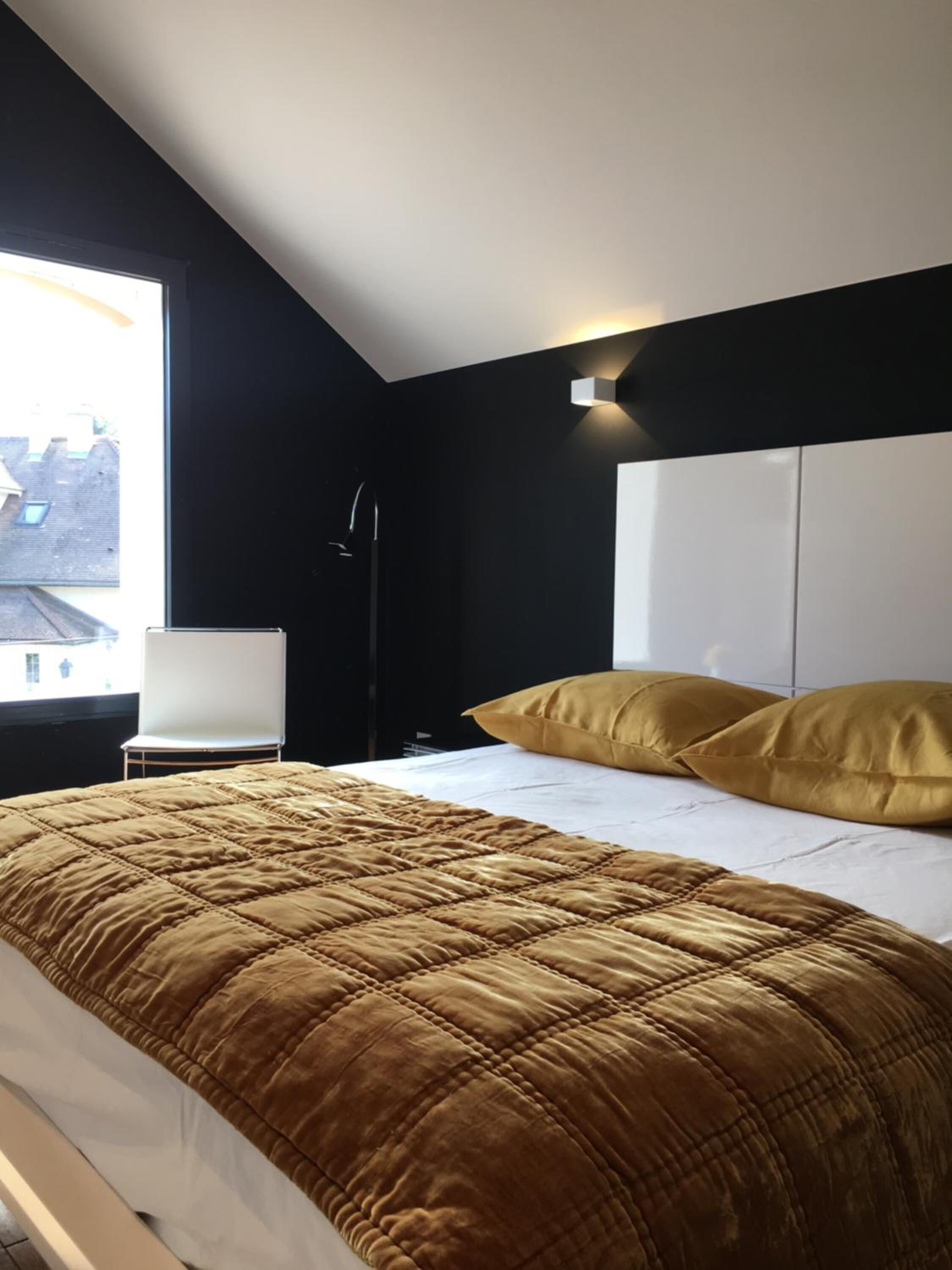 Une chambre moderne avec un grand lit, deux oreillers, une table de chevet et une fenêtre donnant sur des immeubles.
