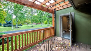Une terrasse en bois avec un toit en verre, une porte donnant sur une salle de bain et une vue sur un parc arboré, avec une route.