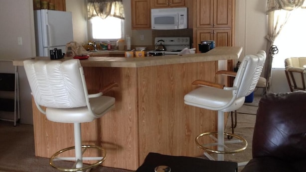 Una cocina con armarios de madera, un refrigerador blanco, un microondas y una estufa. En la zona de estar hay dos taburetes blancos y un sofá de cuero marrón.