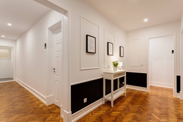 Un couloir aux murs blancs, avec des lambris noirs et un parquet en bois disposé en motif chevron. On y voit des certificats encadrés accrochés au mur, ainsi qu’une petite table sur laquelle est posée une plante.