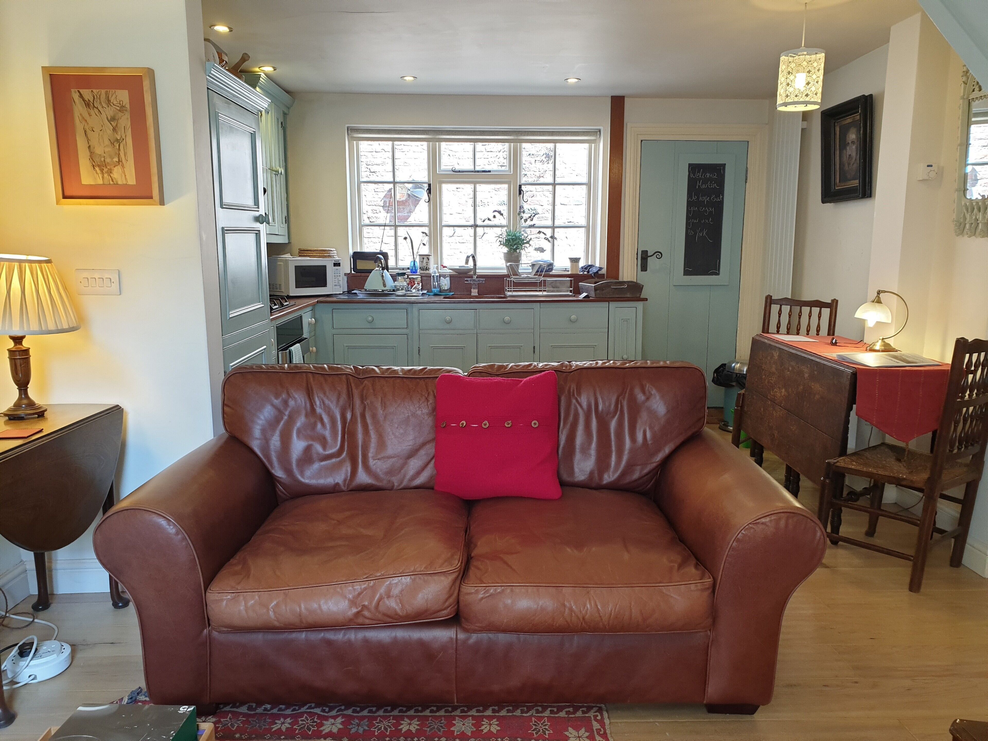 A living room with a brown leather sofa, a dining table with chairs, a kitchen area with cabinets and appliances, and a window with a view of the outside.