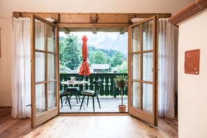 An open sliding glass door leading to a balcony with a table and chairs, a potted plant, and a view of a mountainous landscape.