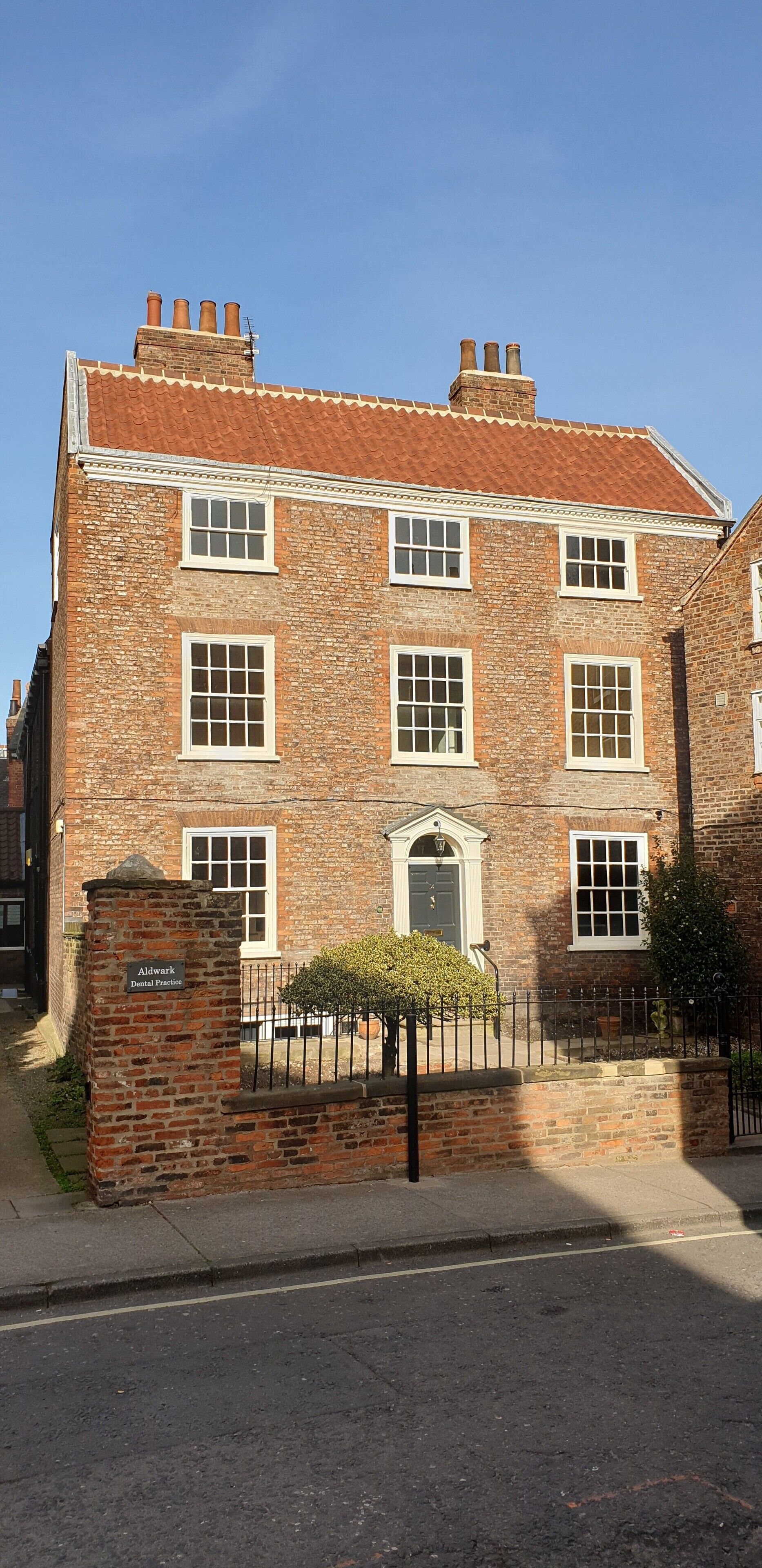 A two-story brick building with a red-tiled roof, a central entrance with a door, and a small garden area with a fence.