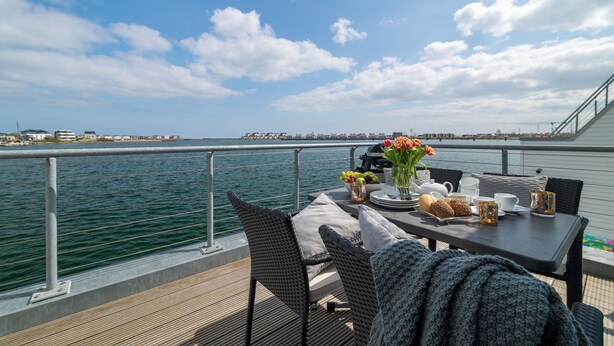 A deck with a table set for dining, overlooking a body of water with a city skyline in the distance. - Water house "Villa Sansibar" - special Baltic Sea vacation with roof terrace and sauna (Olpenitz)