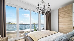 A bedroom with a large window overlooking a body of water, a bed with a patterned blanket, and a chandelier. - Water house "Villa Sansibar" - special Baltic Sea vacation with roof terrace and sauna (Olpenitz)