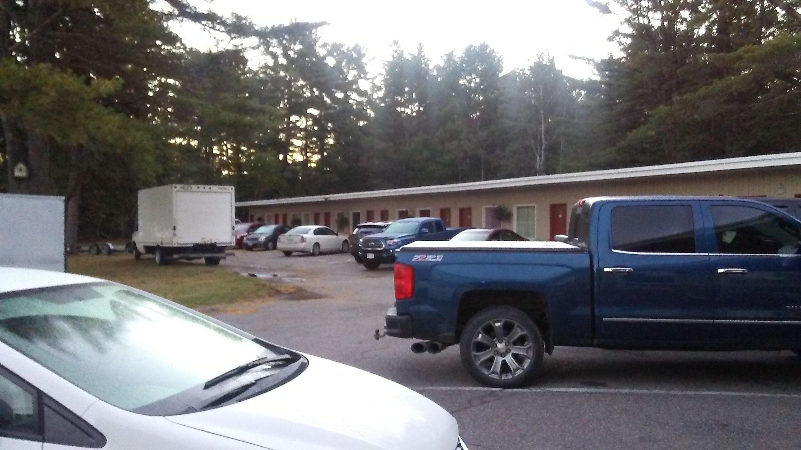 A parking lot with several vehicles, including a white car and a blue pickup truck, in front of a building with multiple rooms.
