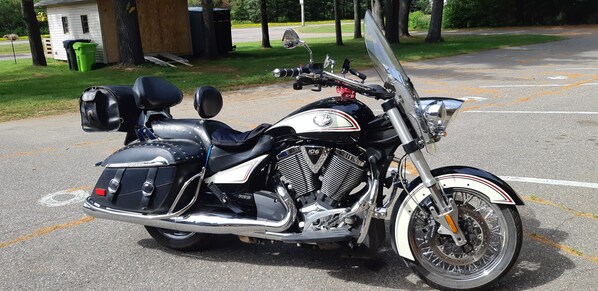 A black Triumph motorcycle parked on a paved surface with a white wall and green trash bins in the background.