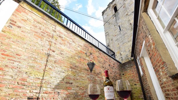 A table with a bottle of red wine and two glasses, set against a brick wall with a metal railing and a stone tower in the background. - 87 Micklegate (York)