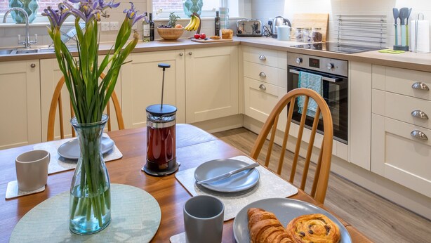 A modern kitchen with white cabinets, a dining table with a vase of flowers, and a plate of croissants. - 71 Micklegate - Apartment 1 (York)