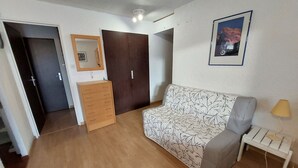 Une pièce avec un plancher en bois, un canapé orné d’un motif à feuilles, une petite table de chevet blanche avec une lampe, une commode en bois, une armoire et un tableau encadré accroché au mur.