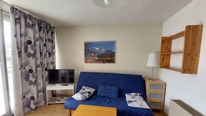 Un salon avec un canapé bleu, une table basse en bois, un téléviseur posé sur un meuble blanc, un tableau représentant des montagnes, une lampe et une étagère en bois.