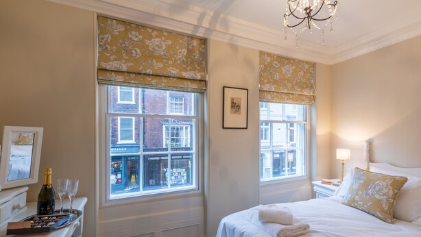 A hotel room with a bed, a bedside table with a bottle and glasses, a window with a view of a building, and a chandelier. - 71 Micklegate - Apartment 3 (York)